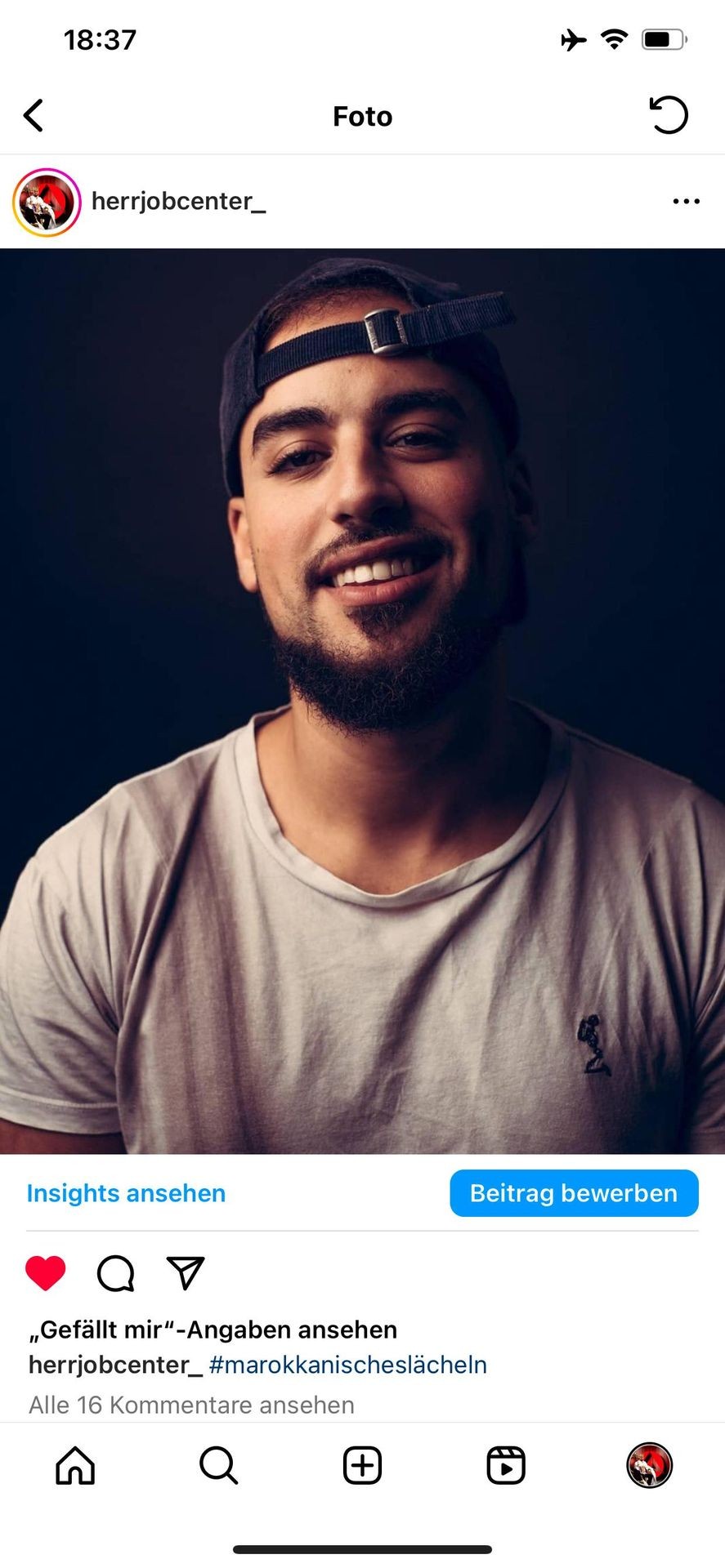 Smiling man wearing a backwards cap and a white t-shirt in a dimly lit setting.