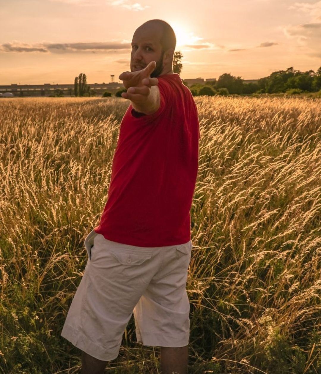 Person in a red shirt pointing towards the camera while standing in a grassy field at sunset.