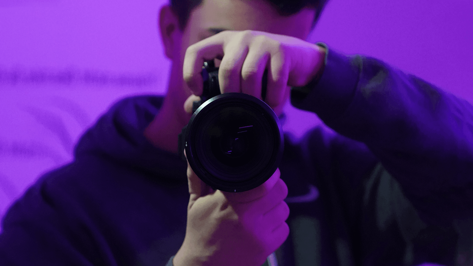 Person holding and pointing a camera towards the viewer, with a purple lighting effect in the background.