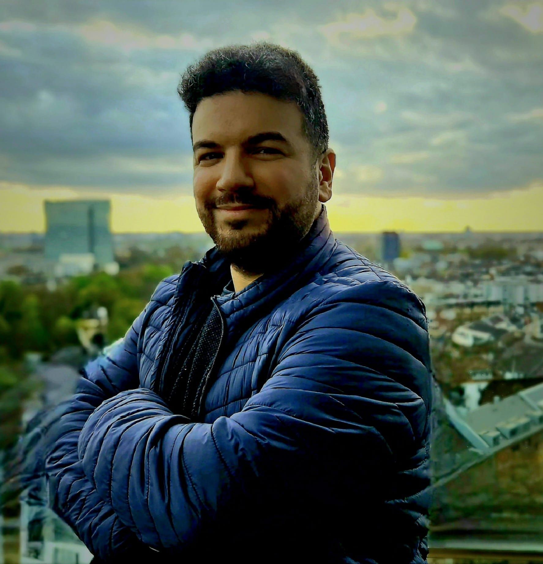 Person in a blue jacket standing with arms crossed, overlooking an urban landscape under a cloudy sky.